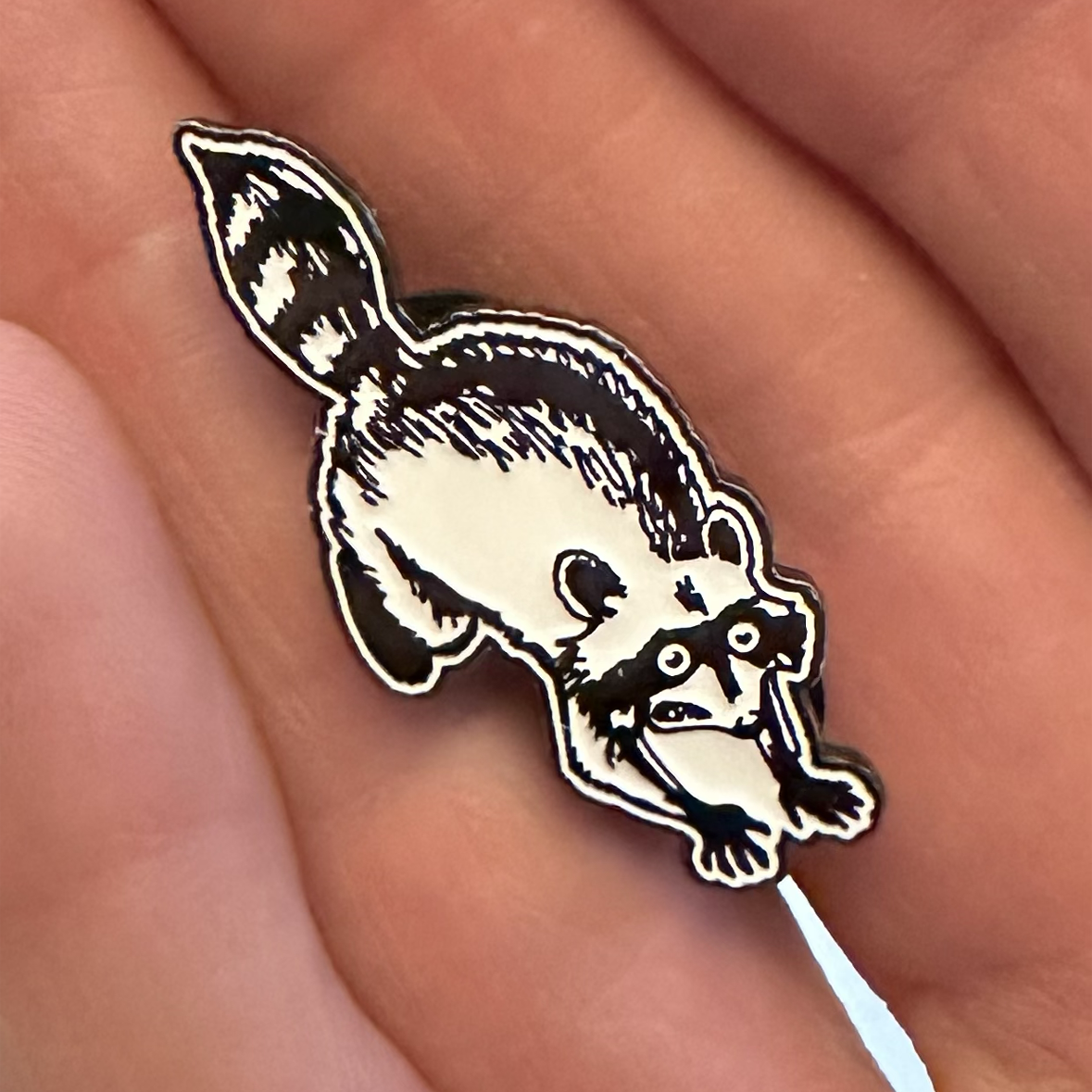 soft enamel pin of a panicking raccoon in black and white held in someone's hand