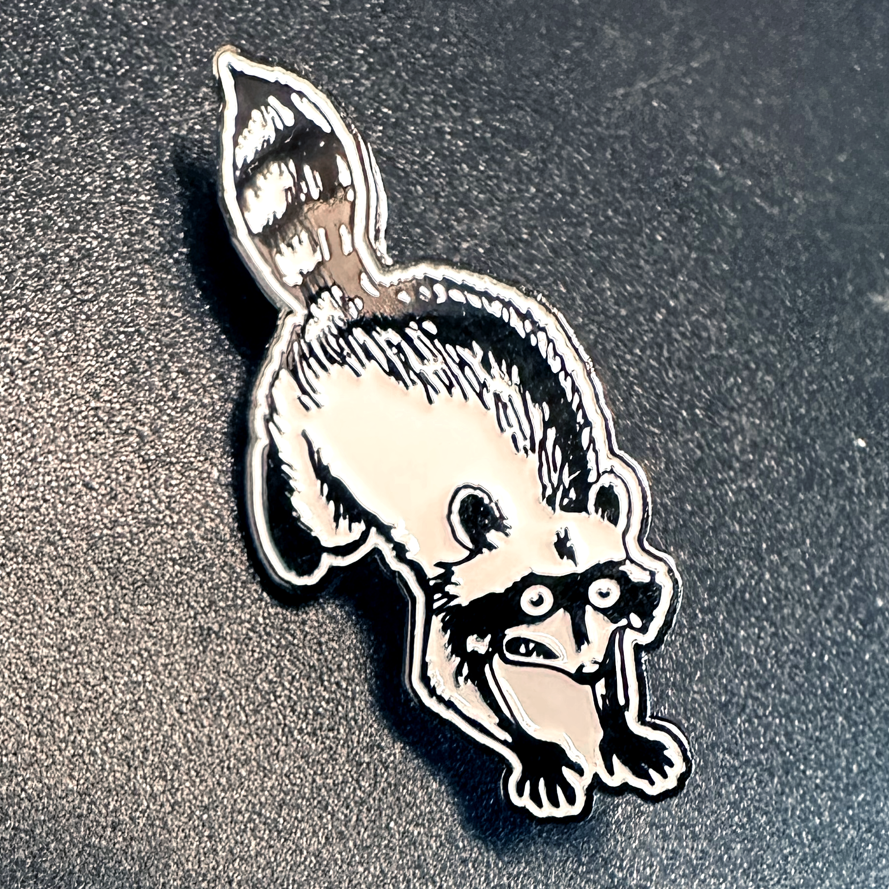 soft enamel pin of a panicking raccoon on a pebbled black surface